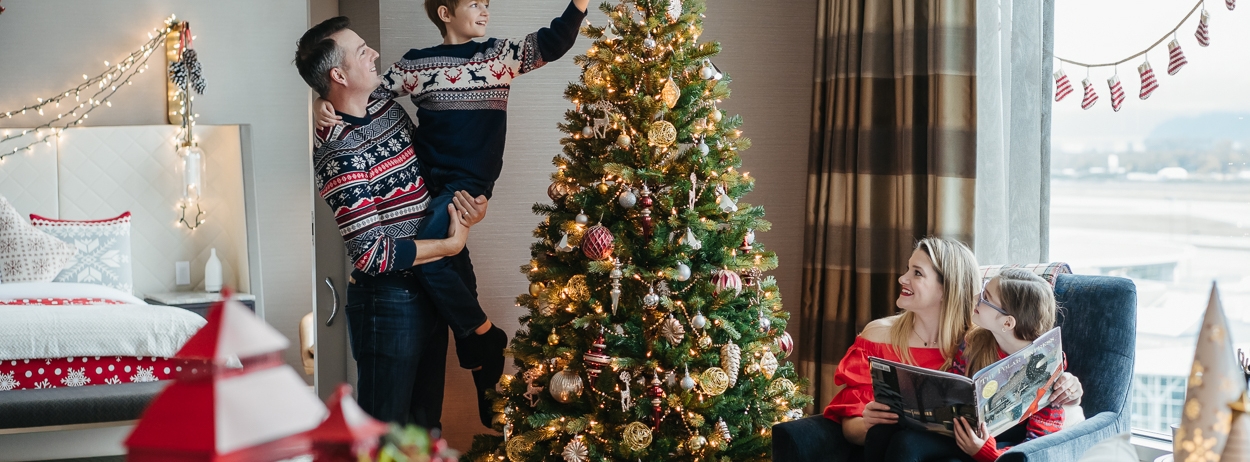 man holding his son while he puts the tree topper on the christmas tree while a mother and daughter read a book