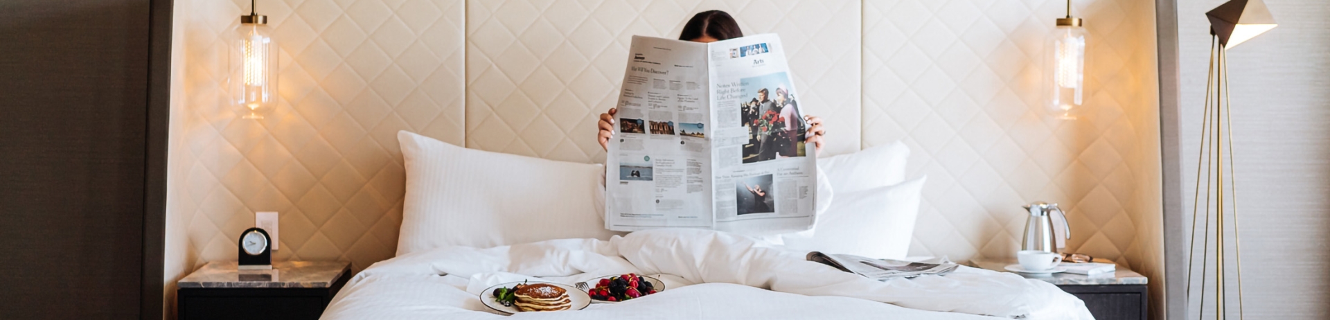 woman reading a newspaper with a tray of breakfast foods at the foot of her bed