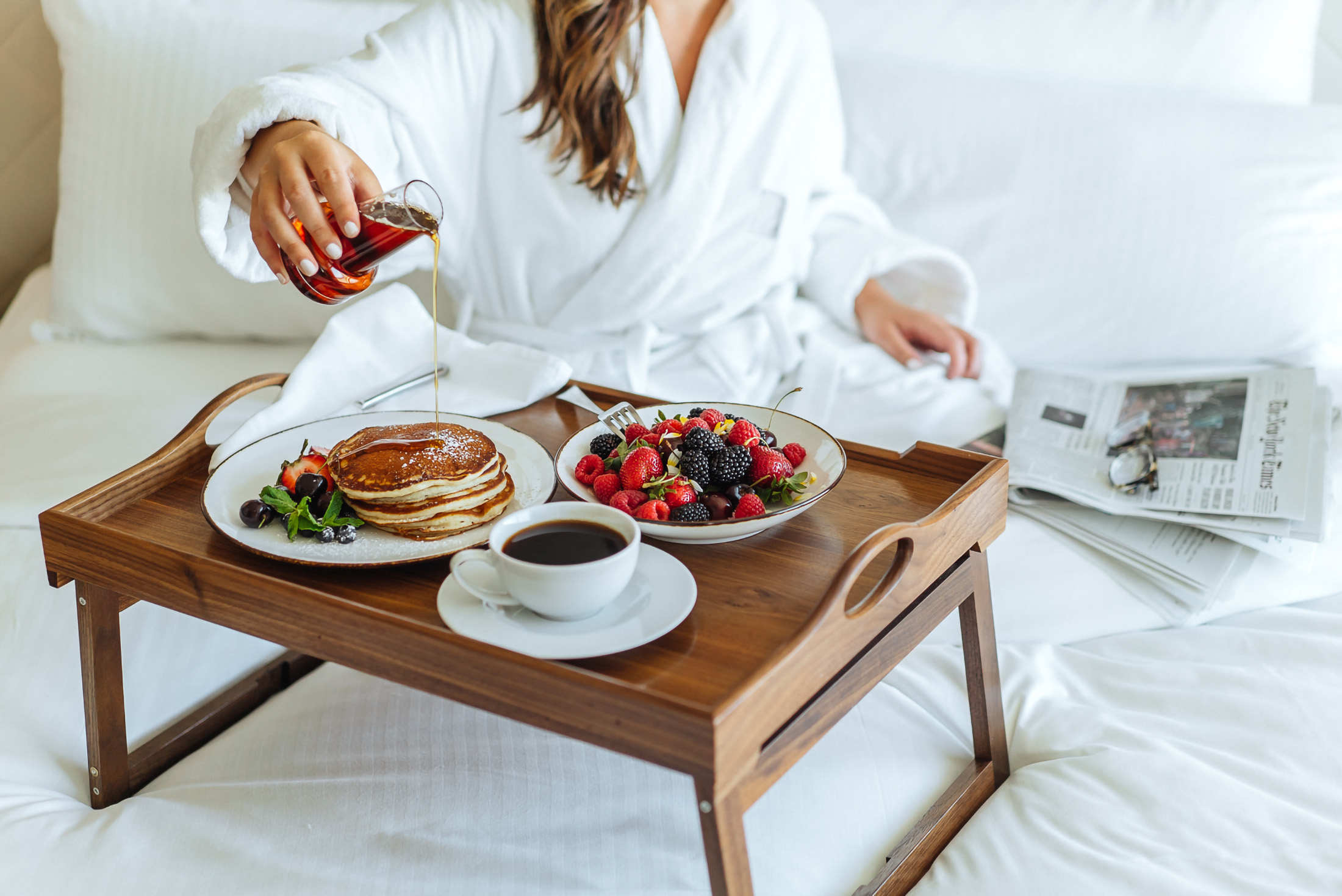 woman pouring syrup on her pancakes while eating her breakfast in bed