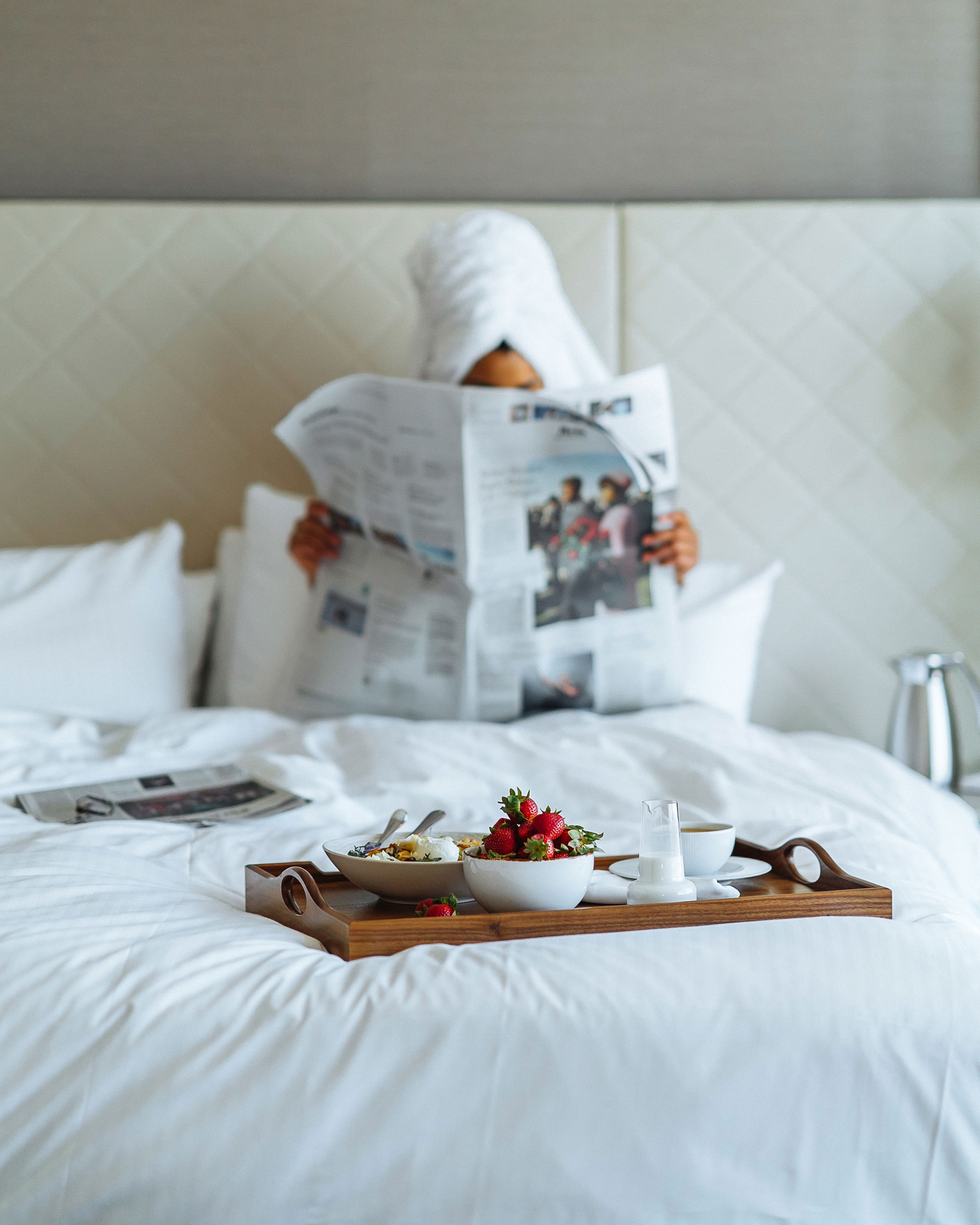 woman reading a news paper with a tray of fruit at the end of the bed