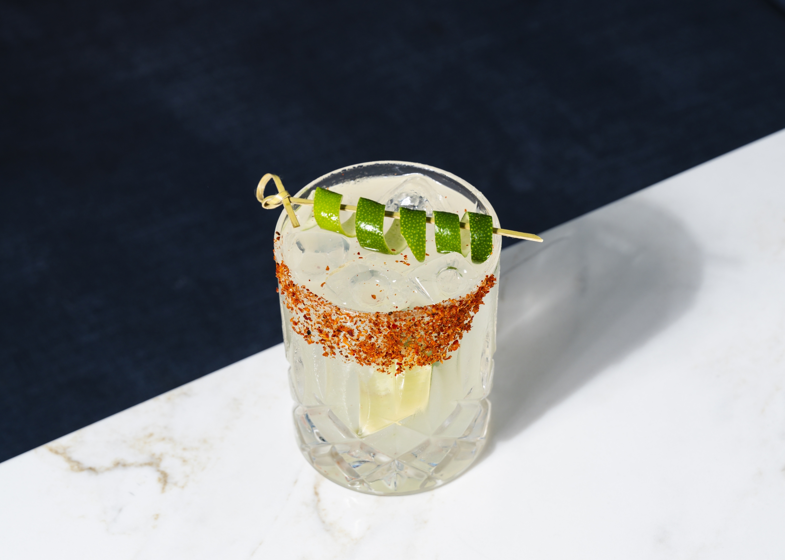 A brightly lit spicy margarita in a crystal lowball glass sits on the edge of a marble table. It has a red tanjin rim and is garnished with a lime peel twist.